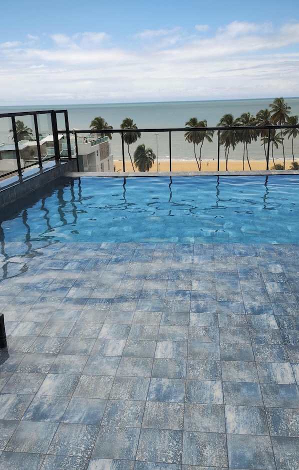 A swimming pool with a tiled deck, a glass railing, and a view of the beach and ocean.
