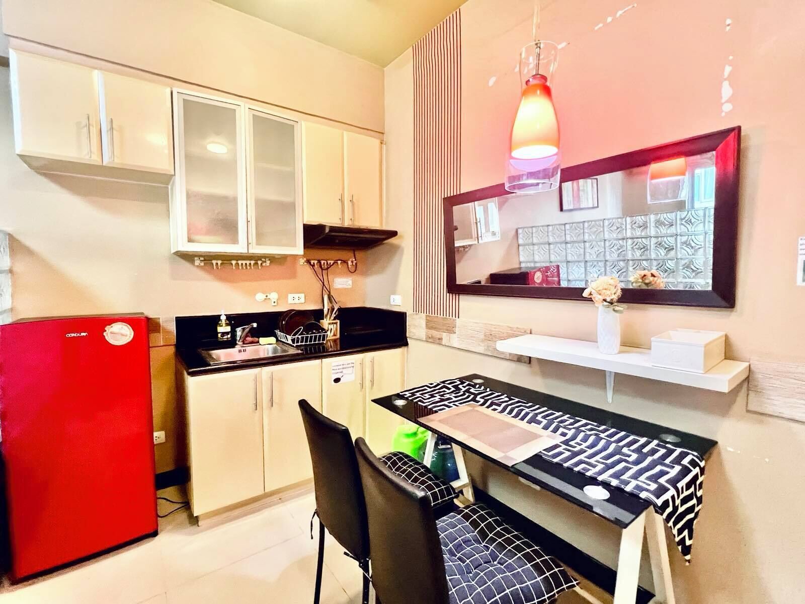A compact kitchen with a red refrigerator, a dining table with a patterned tablecloth, and a hanging pendant light.