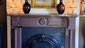 Une cheminée surmontée d’un tableau encadré, de part et d’autre de laquelle se trouvent deux vases ornés de fleurs, et un lustre suspendu au plafond.