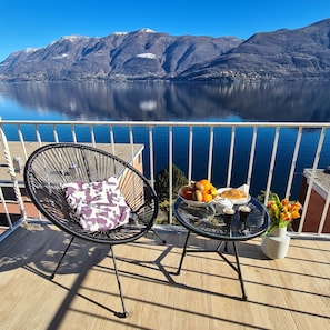 Un balcón con una silla, una mesa con comida y bebidas, y vistas a las montañas y al agua.
