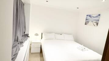 A bedroom with a bed, a nightstand, a lamp, a window with curtains, and a painting on the wall.