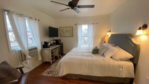 Un dormitorio con cama, ventilador de techo, escritorio con silla, televisor y ventana con cortinas.
