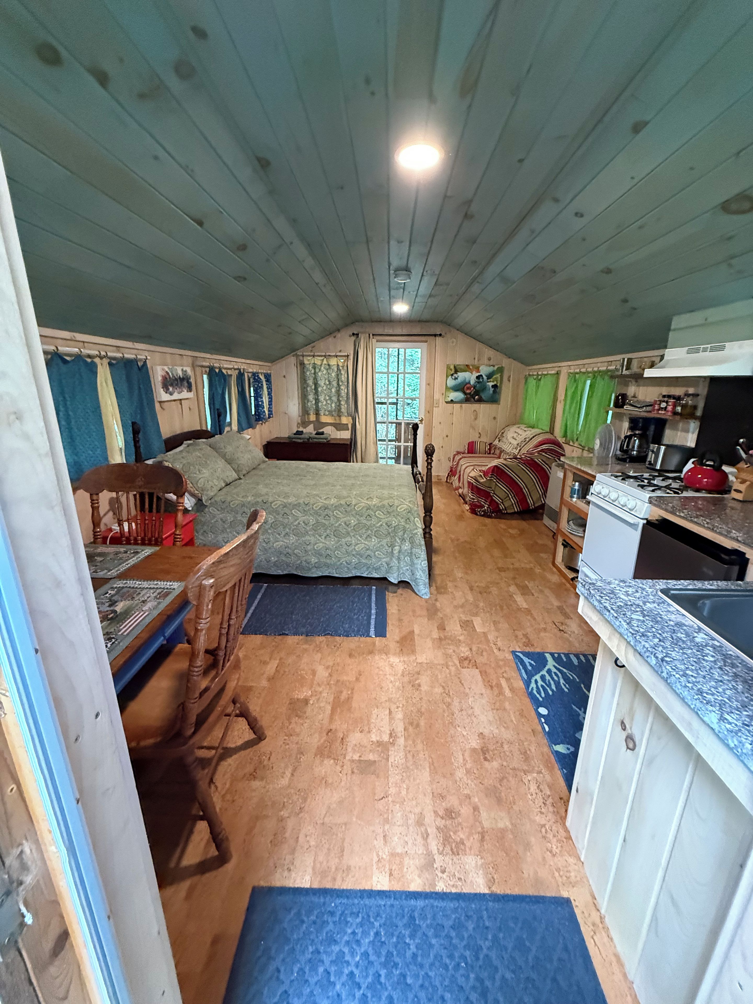 Interior acogedor de una cabaña con cama, una mesa de comedor, una zona de cocina y una ventana con cortinas.