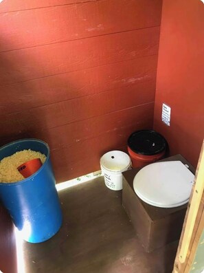 Casa de banho pequena e compacta, com uma parede vermelha, uma sanita branca, um contentor de lixo azul e um recipiente branco com tampa preta.
