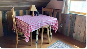 Una habitación acogedora con techo de madera, una mesa cubierta con mantel a cuadros, una lámpara y una ventana con cortinas.