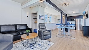 A modern living room with a black leather sofa, a round coffee table, and a gray armchair. A kitchen area is visible in the background with a dining table and bar stools.