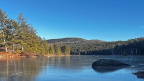 A frozen lake with clear reflections, surrounded by trees and a forested hill in the background.