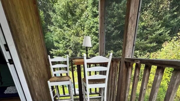 A wooden deck with two white chairs and a small table, a doormat with a mushroom design, and a view of greenery through the railing.
