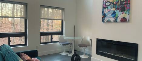 A modern living room with a teal sofa, a fireplace, and a large window overlooking a forest.