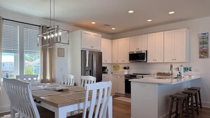 A modern kitchen with white cabinets, a central island, and a dining area with white chairs. - New! Bayfront home w/ Pool, Golf Cart, Kayaks, Bikes & The Islands Best Sunsets! (Saint George Island)