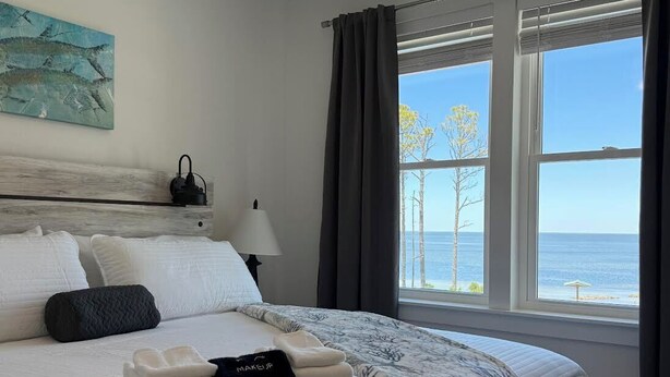 A hotel room with a bed, a lamp, a view of the sea, and a painting on the wall. - New! Bayfront home w/ Pool, Golf Cart, Kayaks, Bikes & The Islands Best Sunsets! (Saint George Island)