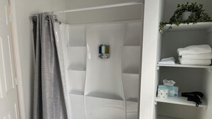A bathroom with a shower, a shelf with towels, a box of tissues, and a hairdryer. - Charming Room 6 in the St. Maries Inn (Saint Maries)