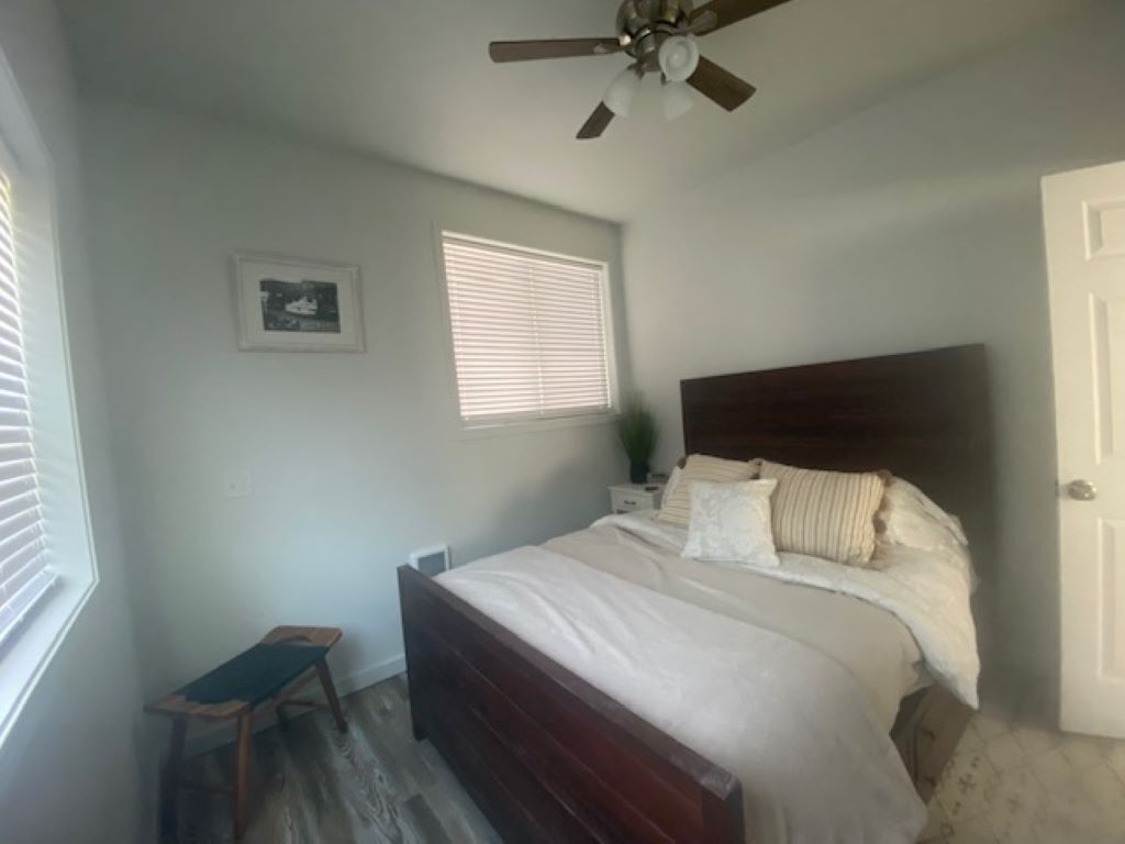A bedroom with a bed, a ceiling fan, a window with blinds, a bedside table, and a small stool.