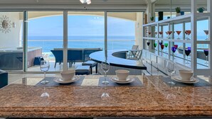 A modern dining area with a view of the sea, featuring a round table, chairs, and a bar area with colorful glassware. - PrivateJacuzzi-LasOlasCondo-Pool&Jacuzzis-Rosarito (Playas de Rosarito)