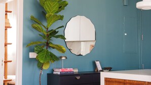 A room with a blue wall, a large plant, a mirror, a wooden shelf, and a desk with books and a lamp.