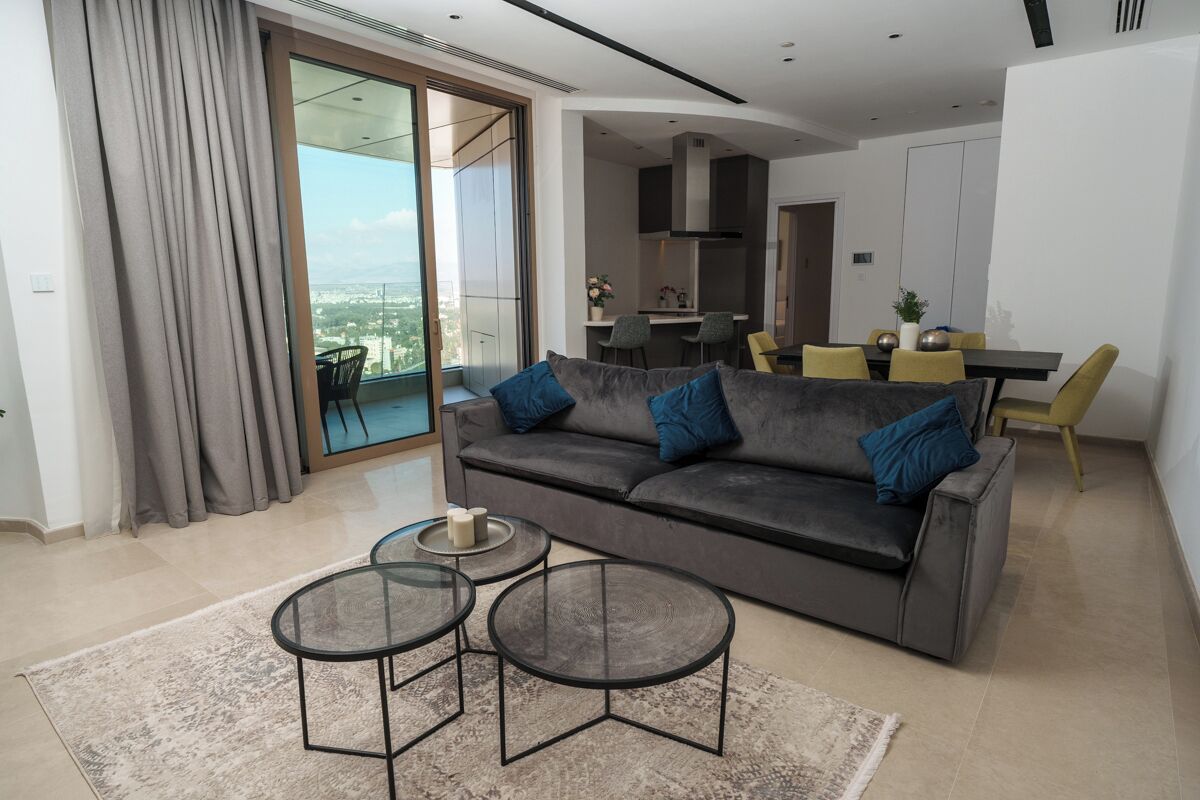 A modern living room with a grey sofa, two round glass tables, and a view of a cityscape through large glass doors.