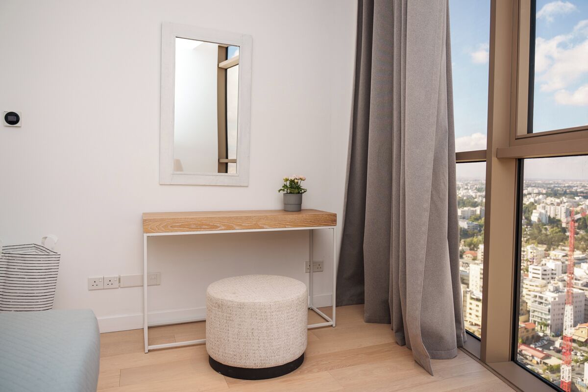 A modern room with a wooden desk, a round ottoman, a mirror, and a view of a cityscape through large windows.