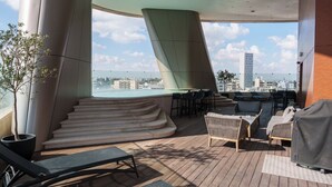 Una terraza en la azotea con vistas a la ciudad, que cuenta con un área de estar con sillas y mesas, una planta en maceta y una escalera que lleva al nivel superior.