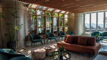 Un espace de détente moderne avec des fauteuils turquoise, un canapé marron et une grande fenêtre donnant sur des palmiers et des immeubles.