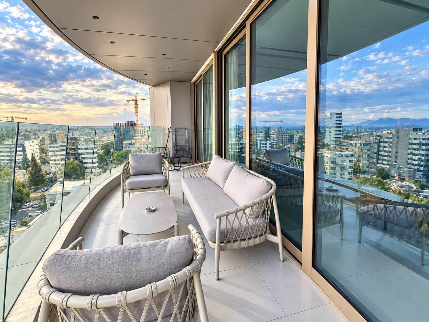 A balcony with a sofa, chairs, and a small table, overlooking a cityscape with buildings and a construction crane.