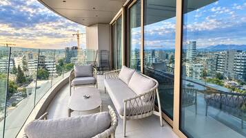 A balcony with a sofa, chairs, and a small table, overlooking a cityscape with buildings and a construction crane.