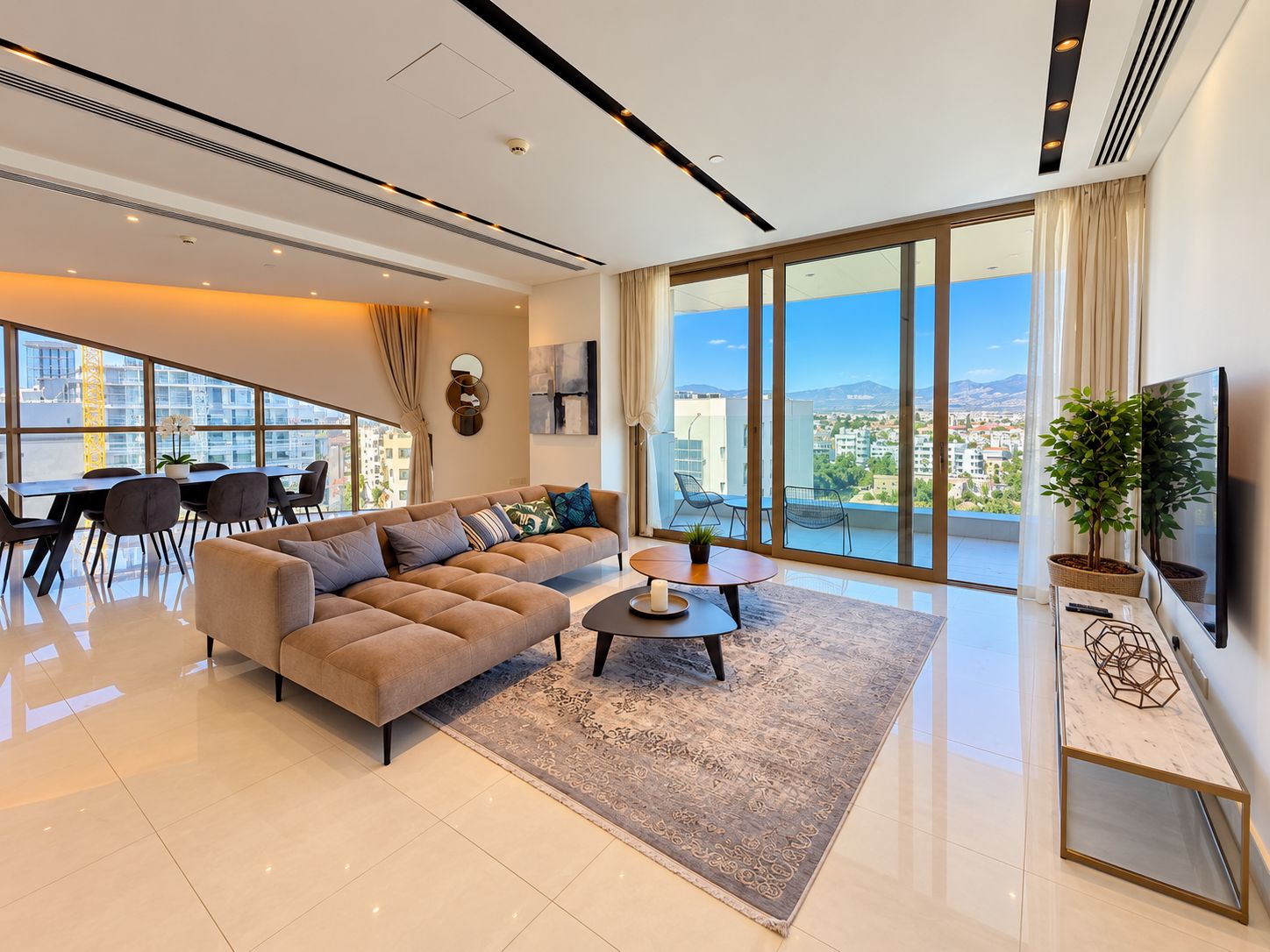 A modern living room with a large sofa, a dining area with chairs, a coffee table, and a TV on a stand.