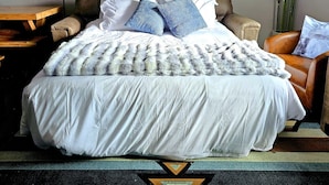 Una cama con ropa de cama blanca, dos almohadas azules y una manta estampada. Hay un cuadro sobre la cama, una planta a la derecha y una mesa de madera a la izquierda.