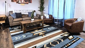 Una sala de estar con sofá, sillón, mesa de centro y un amplio ventanal con cortinas.