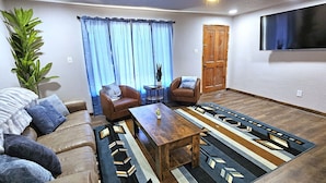 Una sala de estar con un sofá, dos sillones, una mesa de centro, una alfombra, una planta en maceta, una puerta y un televisor montado en la pared.