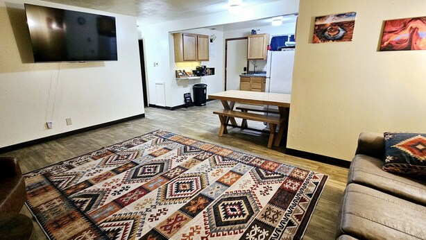 A living room with a patterned rug, a television mounted on the wall, a dining area with a table and chairs, and a kitchen in the background. - 1Bd, King, Brkfst, Large TVs, W/D, Sleeps 4, BBQ (Page)