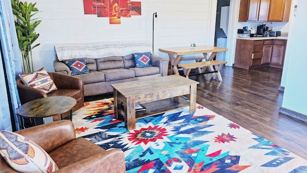 Un salon avec un canapé, une table basse et un tapis à motifs. On aperçoit une cuisine au fond.