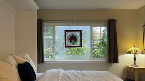 A bedroom with a bed, a window with curtains, a bedside table with a lamp, and a view of greenery outside.