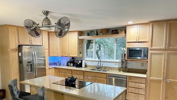 A modern kitchen with wooden cabinets, stainless steel appliances, a central island with seating, and a large window offering a view of the outdoors.
