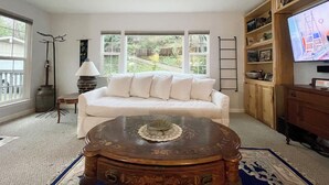 A living room with a white sofa, a wooden coffee table, a TV on a wooden stand, and a bookshelf.