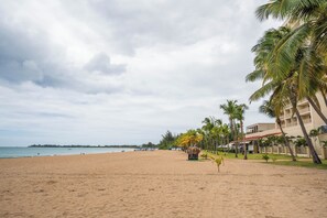 A beach with palm trees, a building with a red roof, and a sign for 'Surfing'. - Beachfront Wave at Isla Verde (Carolina)