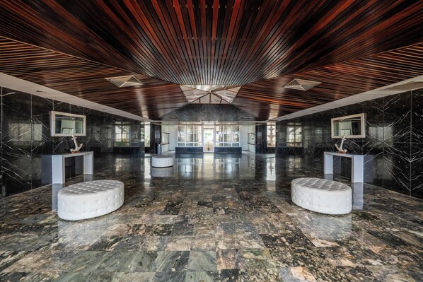 A spacious lobby with a polished marble floor, two white cushioned seating areas, and a high ceiling with wooden slats.