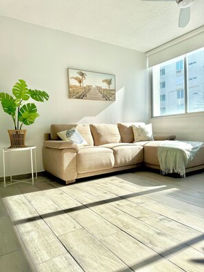 A living room with a beige sofa, a potted plant, a small white table, and a painting of a beach scene.