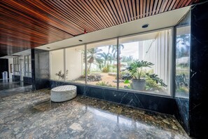 A modern lobby with a marble floor, a white cushioned bench, and a view of an outdoor garden through large windows. - Beachfront Wave at Isla Verde (Carolina)