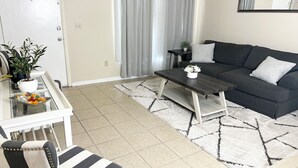 Ein modernes Wohnzimmer mit einer Couch, einem Couchtisch und einem gestreiften Sessel. Eine Topfpflanze und ein kleiner Tisch mit einer Vase stehen links.