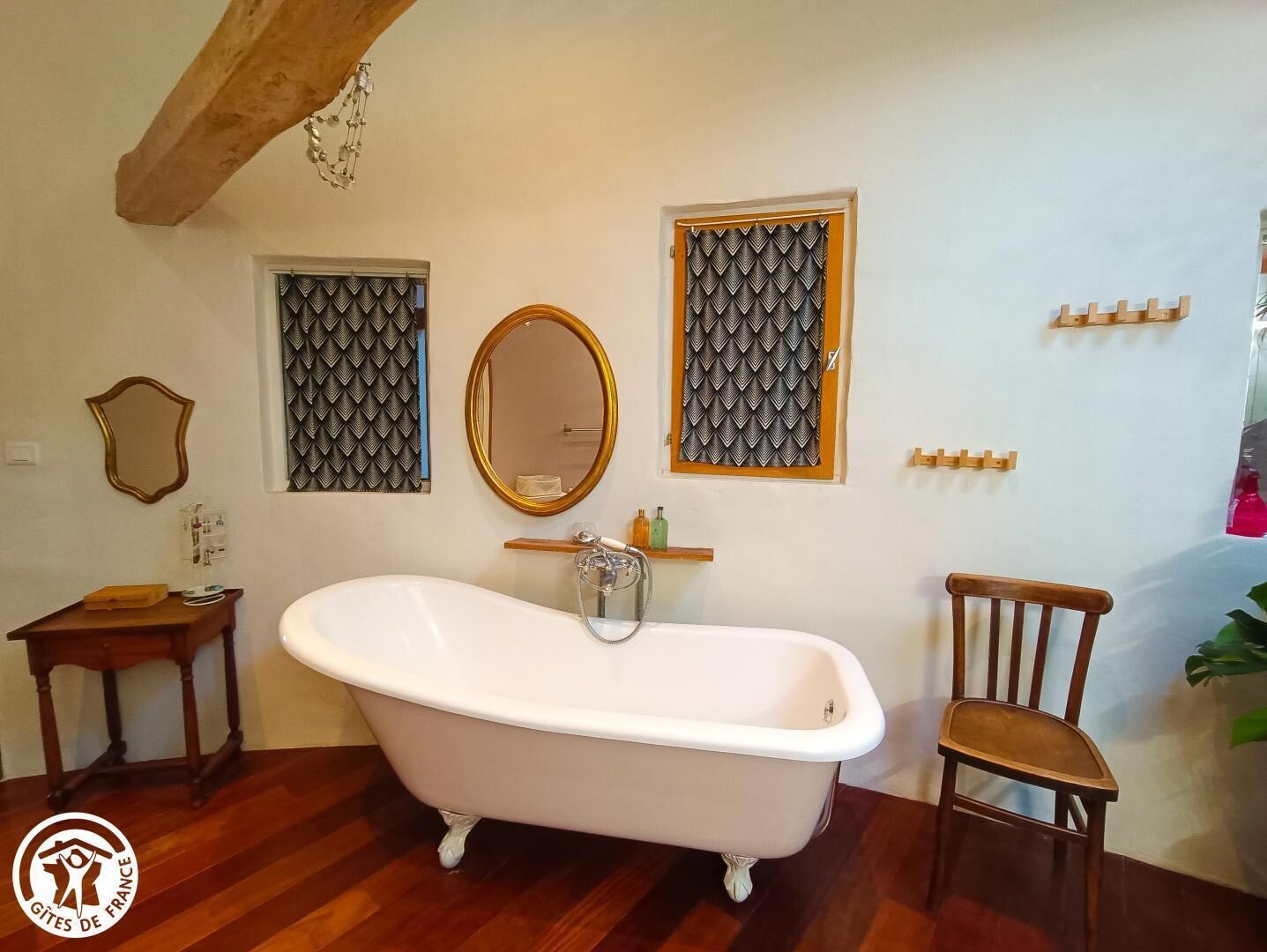 A bathroom with a freestanding bathtub, a wooden chair, a wooden side table, a mirror, and patterned window treatments.