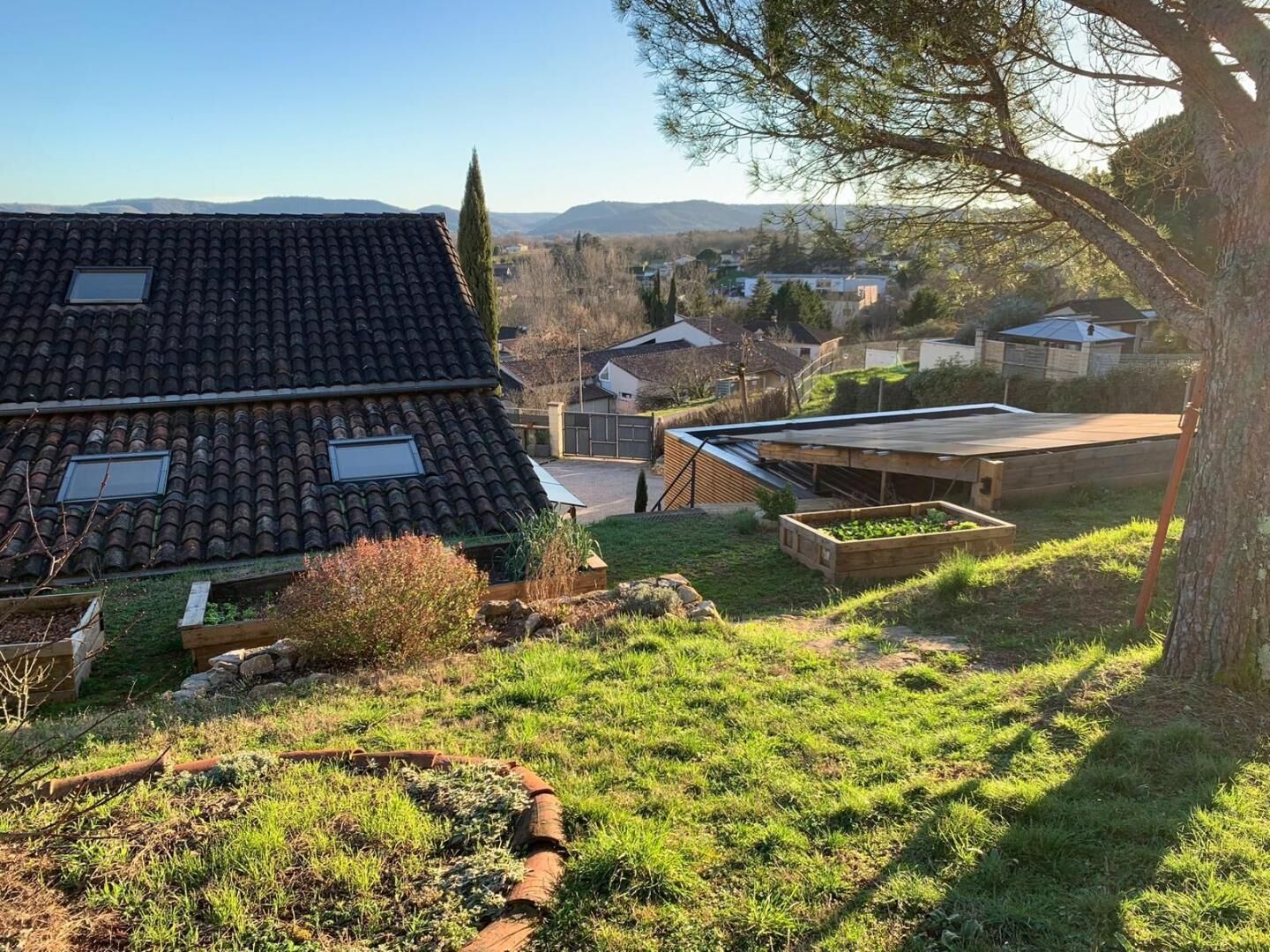 A residential property with a sloped roof, a garden area with raised beds, and a view of a hilly landscape in the background.