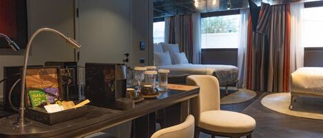 Una habitación de hotel moderna con una cama grande, una zona de estar con una mesa y sillas, una cafetera y una ventana con cortinas.