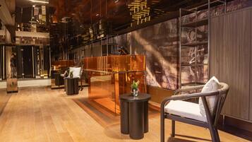 A modern lobby with a wooden floor, a glass reception desk, a seating area with a chair and a small table, and a wall with a large abstract painting.