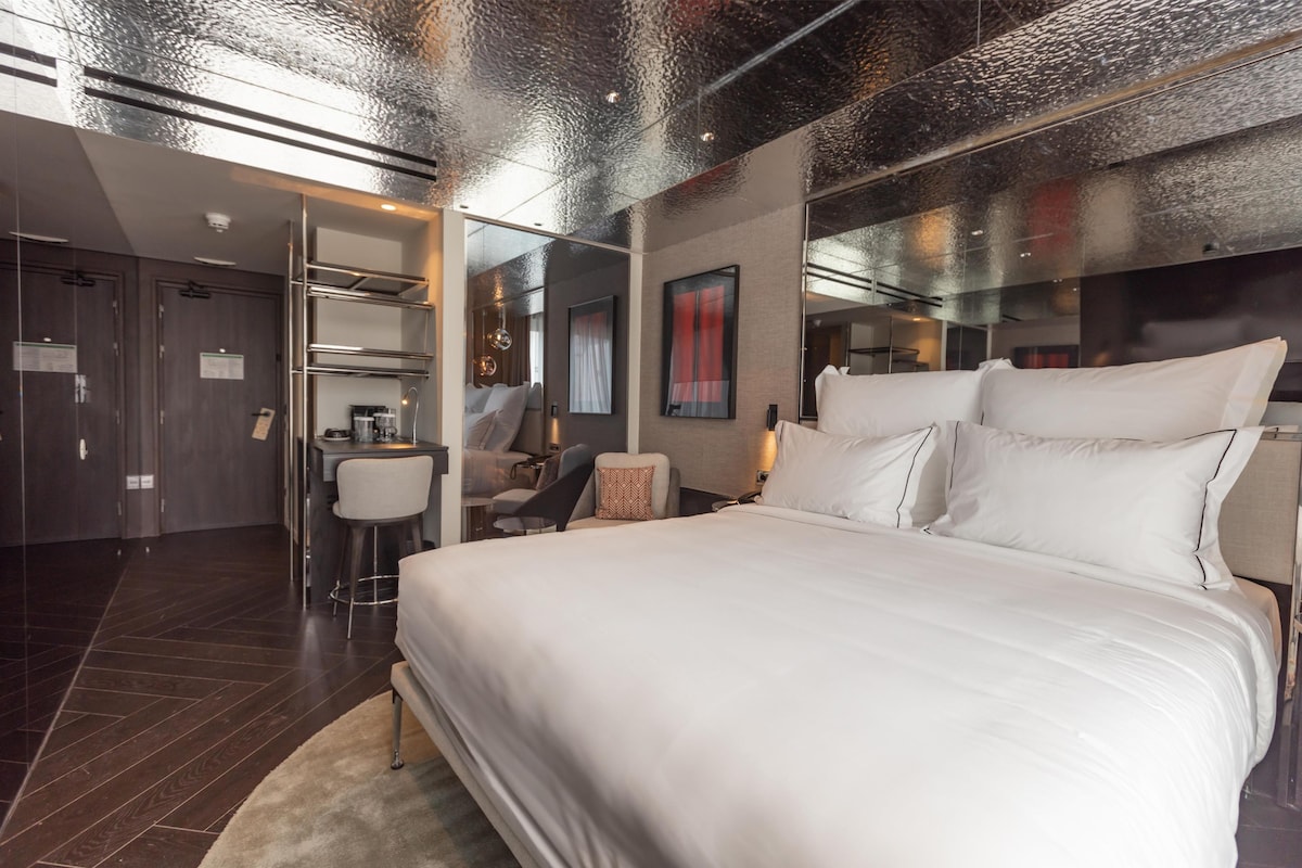 Una habitación de hotel moderna con una cama grande, un escritorio con silla, un pequeño rincón de estar y un baño con ducha e inodoro.