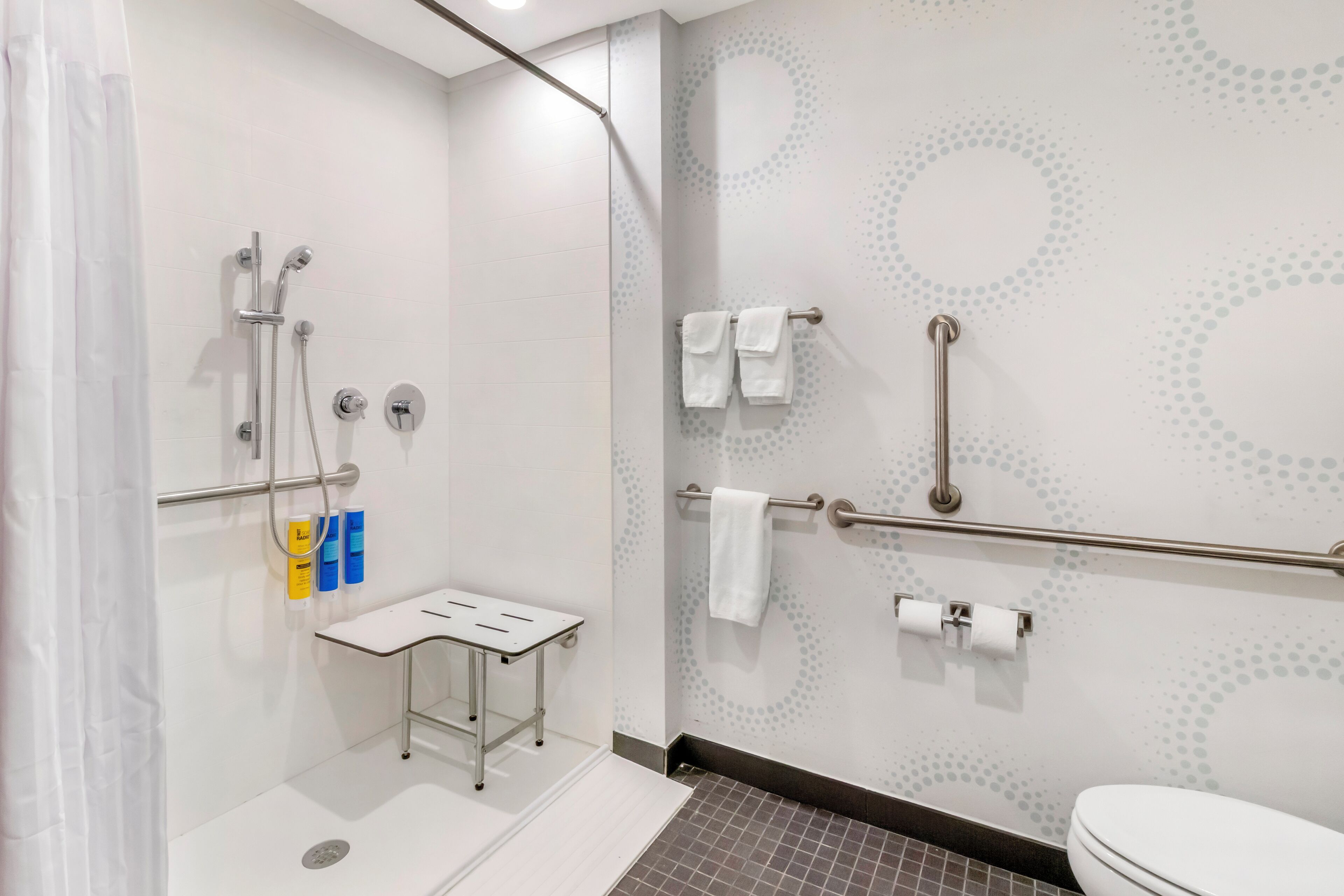 Un baño moderno con ducha, barras de apoyo, inodoro y una pequeña mesa con artículos de aseo.