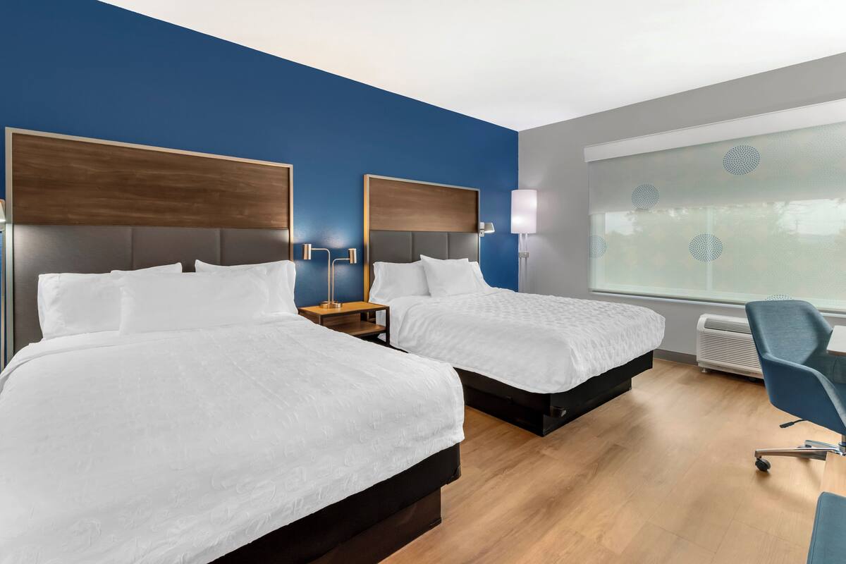 A hotel room with two beds, a desk, a chair, and a large window with blinds.
