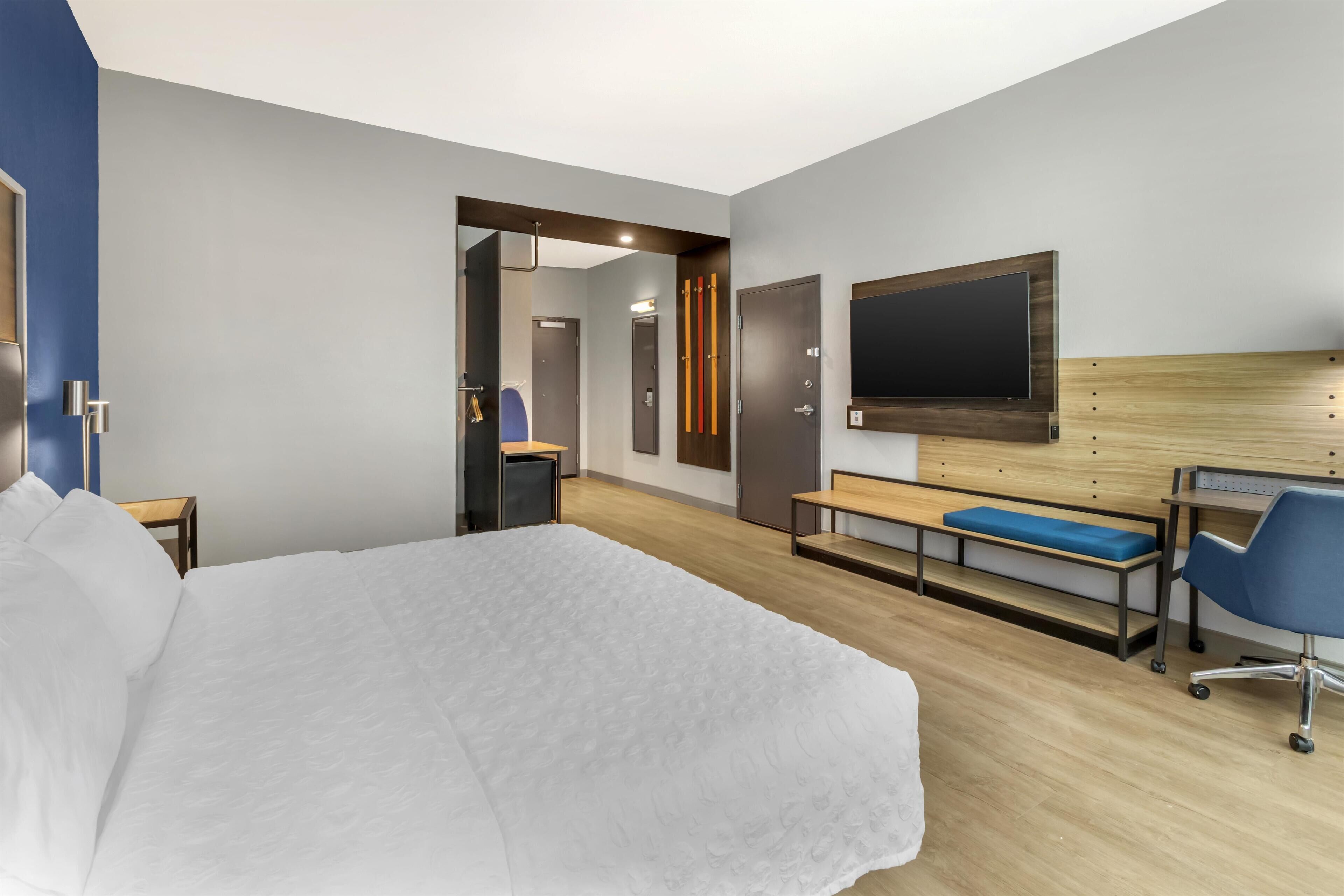 Ein modernes Hotelzimmer mit einem großen Bett, einem Schreibtisch mit Stuhl, einem an der Wand befestigten Fernseher und einem durch eine offene Tür einsehbaren Badezimmer.