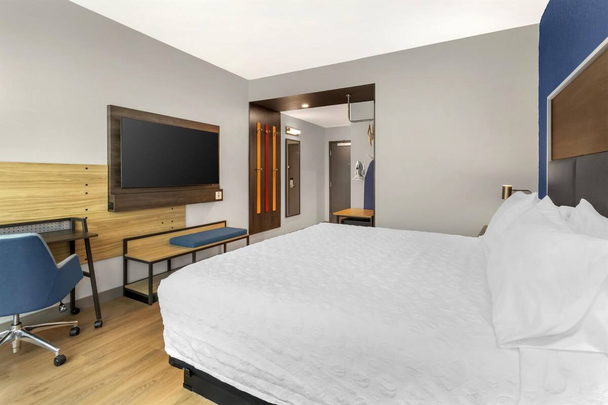 A modern hotel room with a large bed, a desk with a chair, a flat-screen TV mounted on the wall, and a mirror.