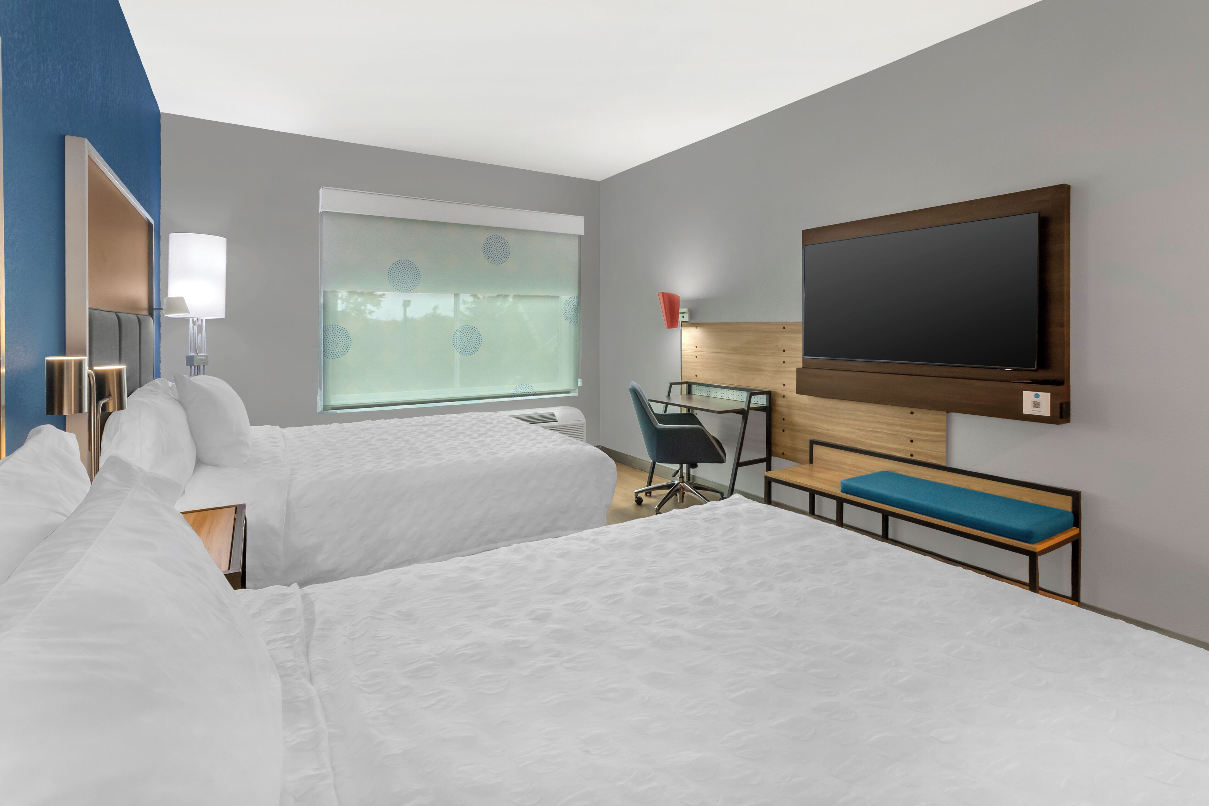 A hotel room with two beds, a desk, a chair, a TV, and a window with blinds.