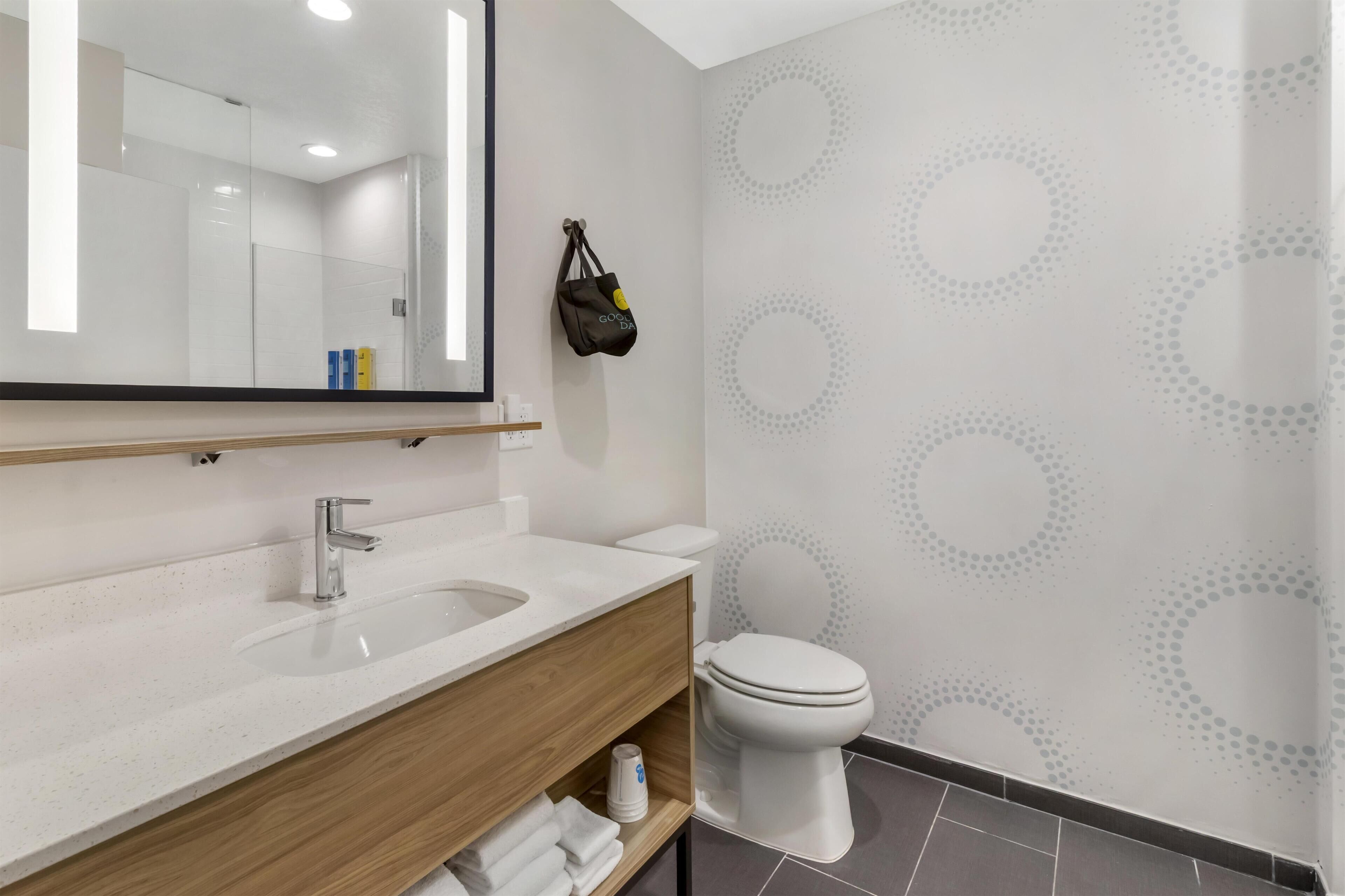 Un baño moderno con un lavamanos blanco, tocador de madera y un inodoro. Un espejo refleja la habitación, y un bolso cuelga en la pared.